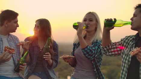 Two-loving-couples-drinks-beer-and-eats-hot-pizza-on-the-roof.-They-are-smile-and-enjoy-the-time-at-the-sunset.-Girl-modestly-looks-at-her-boyfriend